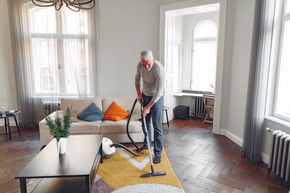 A woman with curly hair wearing a light green jacket, blue jeans, and white sneakers is performing surface cleaning on a patterned area rug in a living room. She is using a yellow vacuum cleaner with 