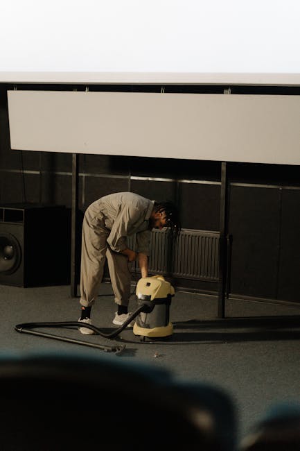 A child wearing beige sneakers, blue jeans, and a beige jacket is using a black vacuum cleaner with a yellow canister to clean a patterned area rug in a residential setting. The rug features floral mo